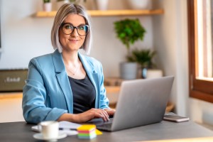 A smiling woman wearing glasses and a blue blazer, sitting in front of a laptop.