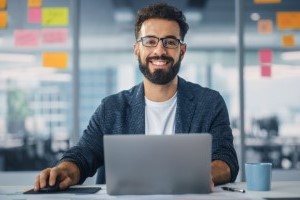 A smiling, young, male person with a beard and glasses. He's sitting in an office space in front of a laptop.